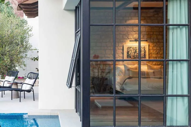 View of a modern glass-walled room from the outside, featuring a cozy bedroom and adjoining outdoor patio with pool, designed by B & L Glass in Santa Rosa.