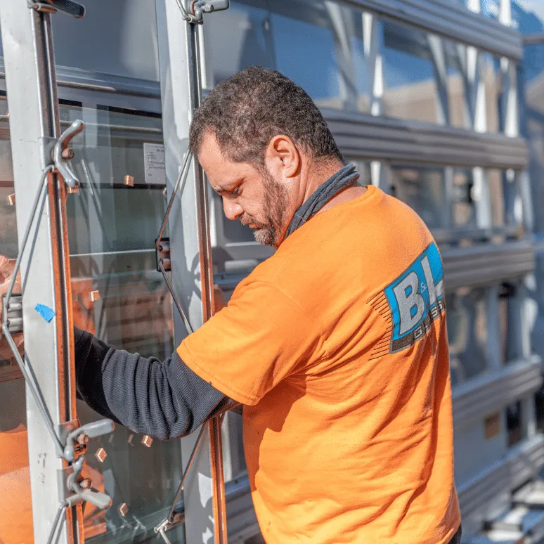 A technician wearing a B & L Glass shirt works on a glass unit, focusing intently on the task, with tools and equipment visible in the background.