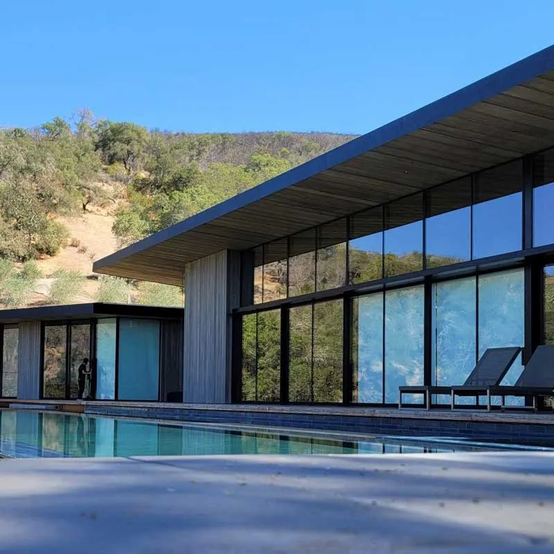 A modern house with large glass windows reflecting trees and hills, featuring a pool and minimalist loungers at the forefront, set under a clear blue sky.