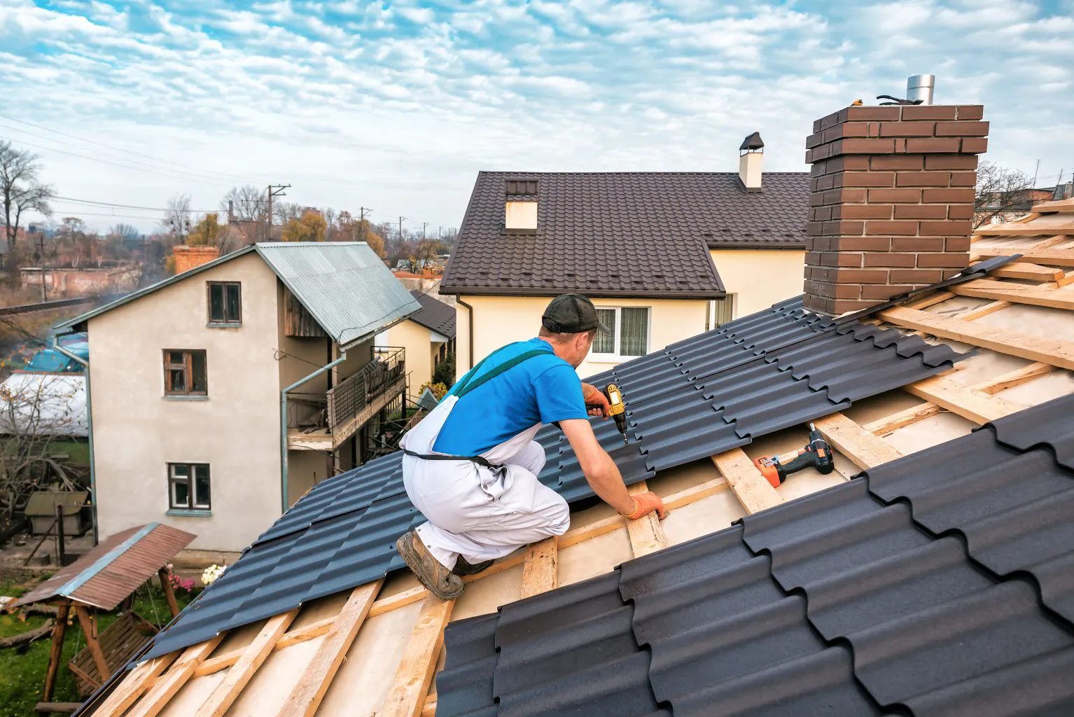 A worker from Bay 101 Roofing, Inc. installs metal sheets on a roof in Alviso, using a drill, surrounded by neighboring houses.