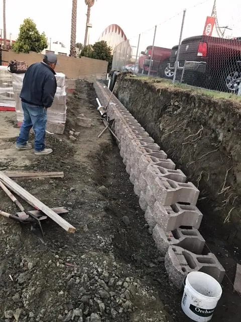 A worker from Bay Area Drainage, Inc. in Moraga is constructing a retaining wall using concrete blocks, with tools and materials visible around the worksite.