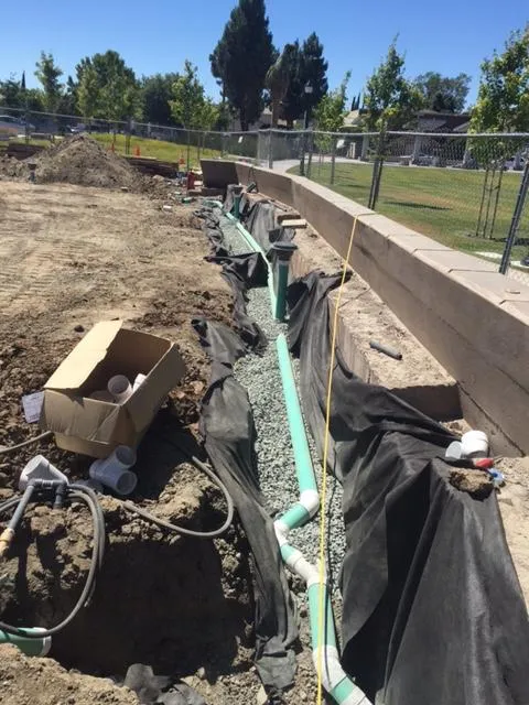 Construction site in Moraga showing drainage pipes being installed by Bay Area Drainage, Inc., with gravel and black fabric lining a trench.
