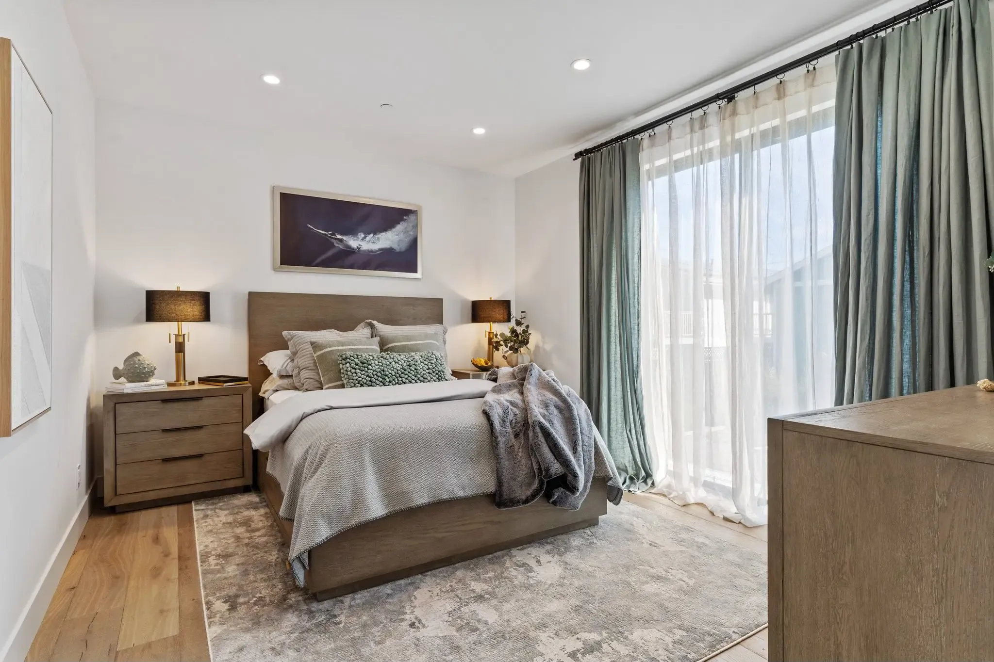 A modern bedroom featuring a wooden bed with grey bedding, flanked by wooden nightstands and lamps, with a large window and full-length curtains.