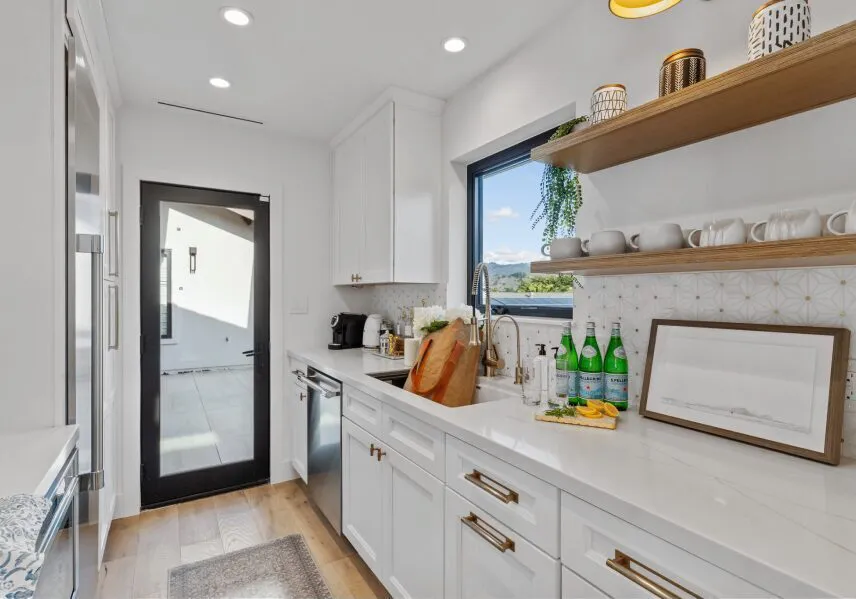 A modern kitchen with white cabinets, a black-framed glass door, and wooden shelves, featuring elegant decor and a scenic view outside the window.