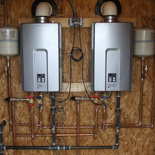 Two modern water heaters installed on a wooden wall, connected by copper piping, with expansion tanks above each unit.