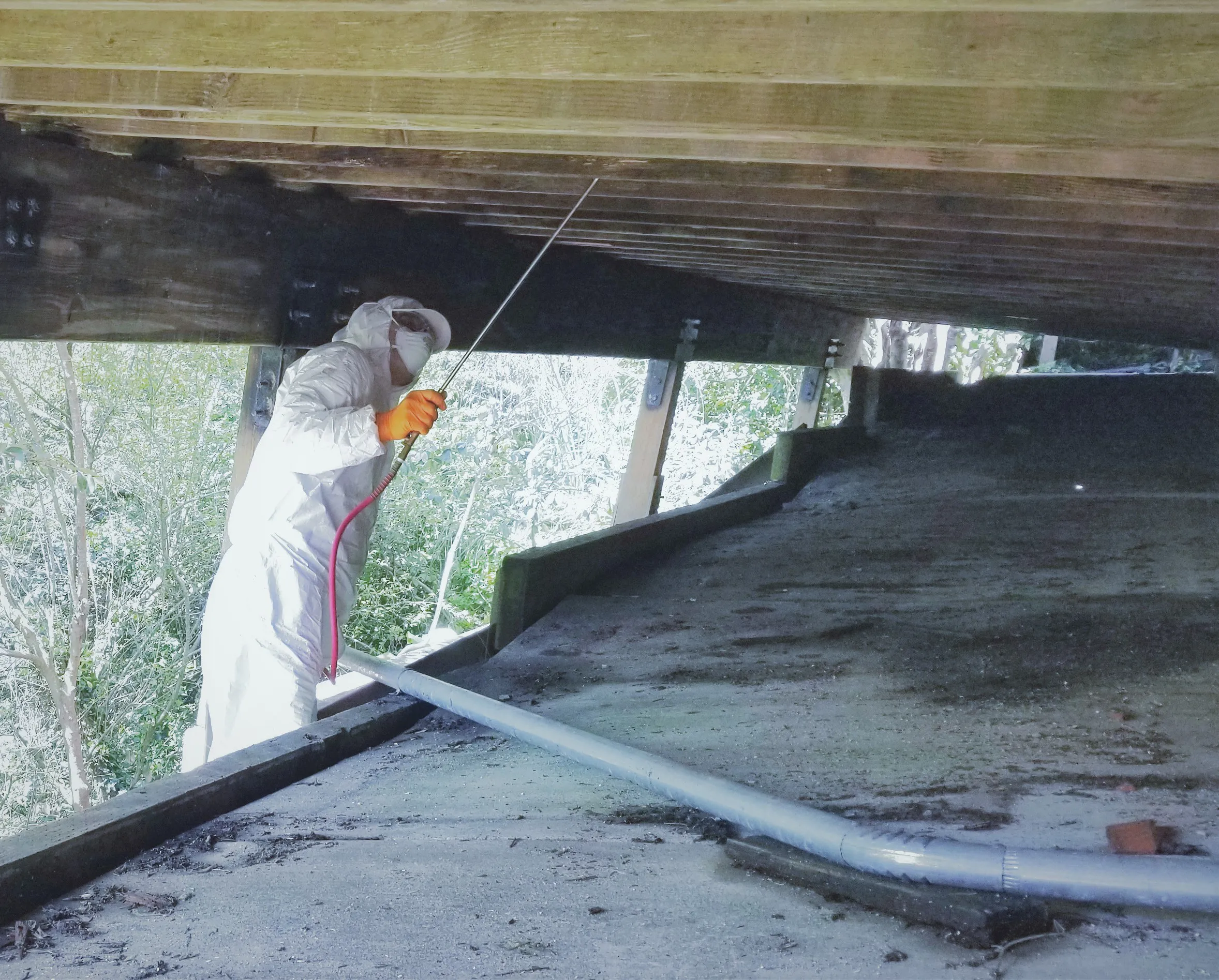 A person in protective gear sprays treatment under a house's wooden structure for termite prevention.