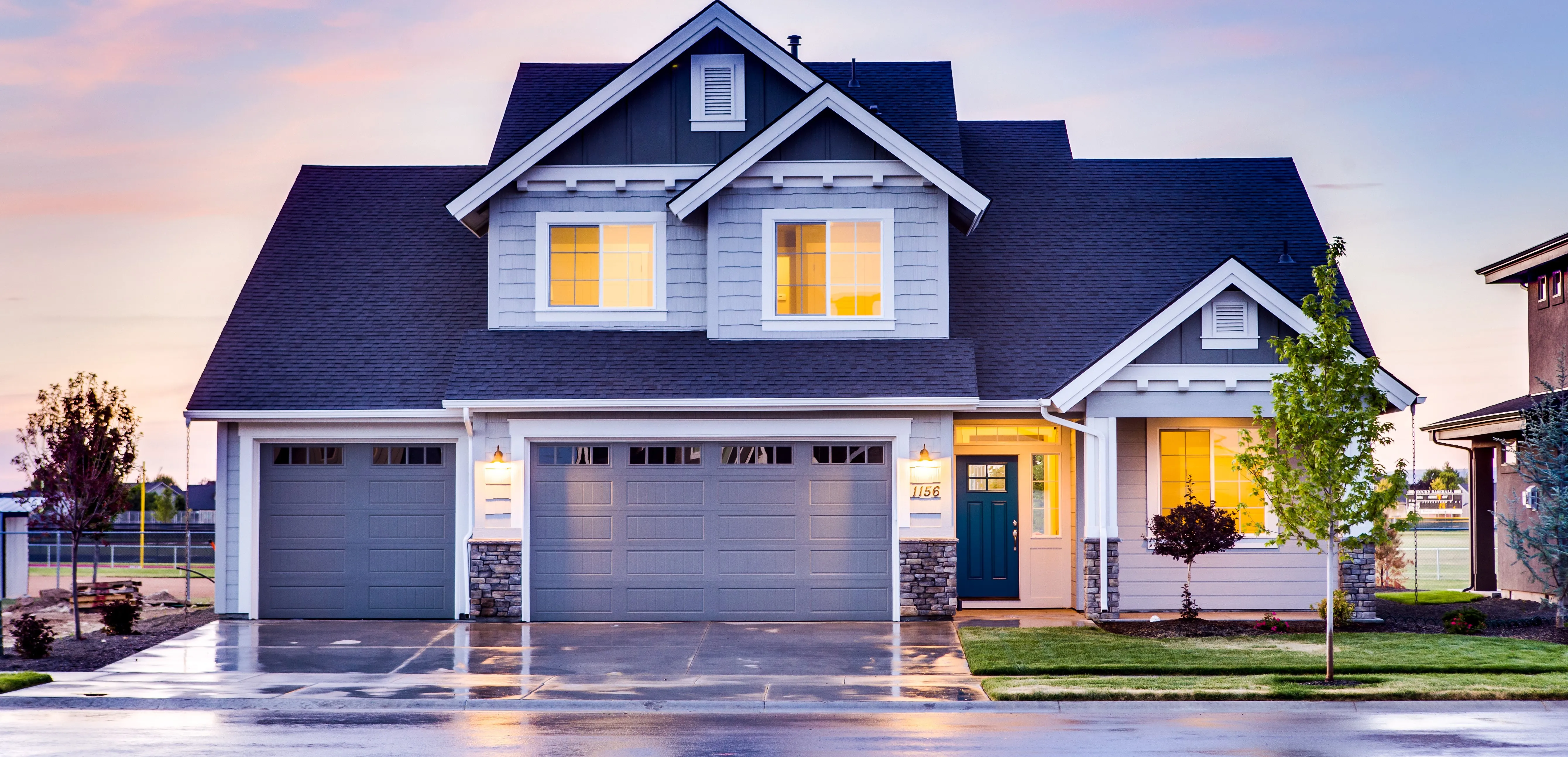 Modern suburban house with a two-car garage at dusk, featuring illuminated windows and a well-kept lawn, suggesting a serene residential area.
