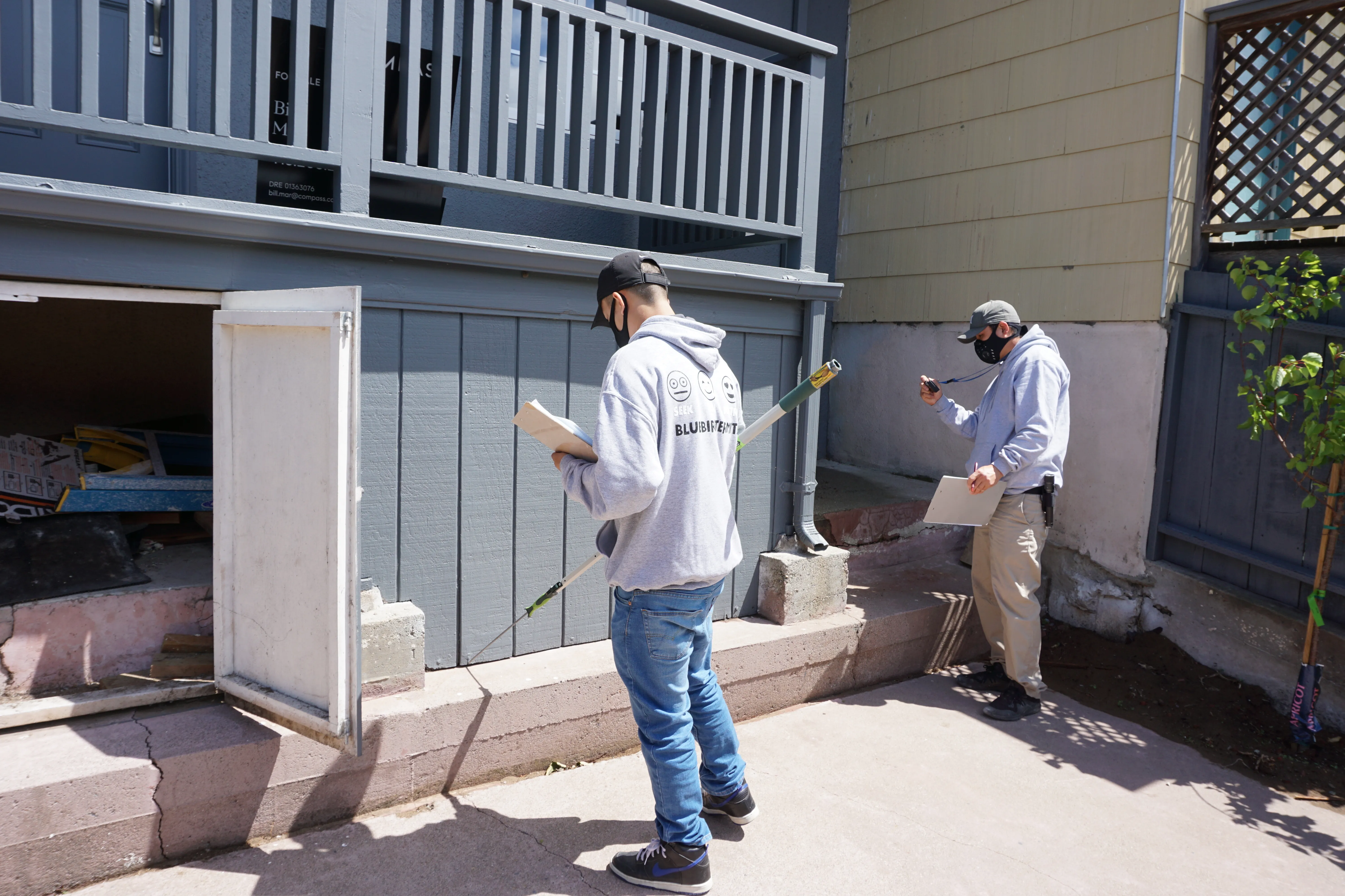 Two Bluebird Termite inspectors in casual attire are conducting a detailed inspection of a property in Burlingame. They are focused on potential issues around a house entrance.