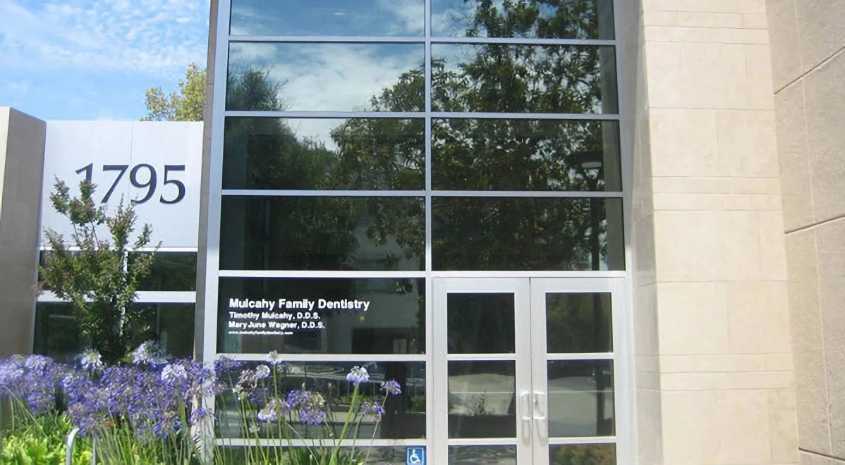 Front of a modern building with large glass windows and entrance doors, showcasing reflective surfaces and landscaping in front.