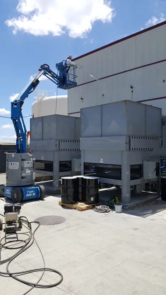 An industrial worker on a lift spray-coats a large facility exterior, with equipment and barrels nearby at CG Coatings Inc in Fairfield.