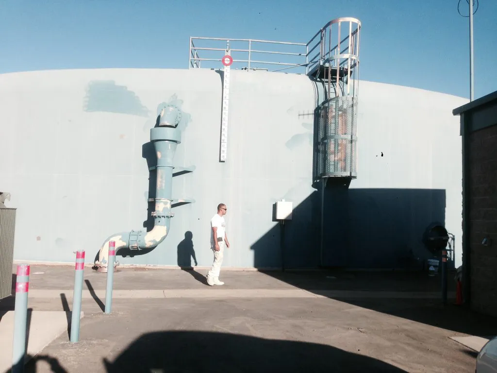 An industrial worker walks past a large, blue-painted storage tank featuring piping and a platform, under a clear sky.