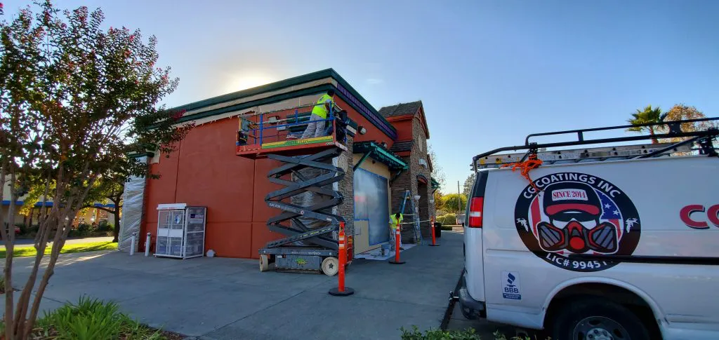 A construction worker stands on a scissor lift applying coatings to the exterior wall of a building in Fairfield, with a CG Coatings Inc van parked nearby.