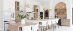 A modern kitchen with light wooden cabinetry, a large island with white chairs, pendant lighting, and built-in shelves, reflecting a sleek and functional design.