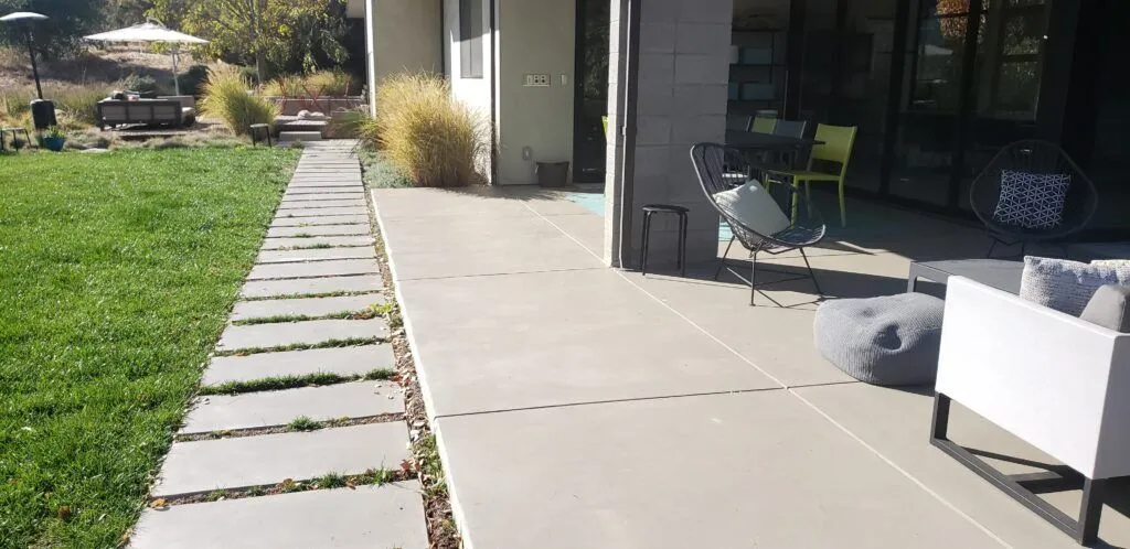 A modern concrete patio in Martinez with minimalist outdoor furniture and a pathway leading through a grassy area, installed by California Concrete Overlay Inc.