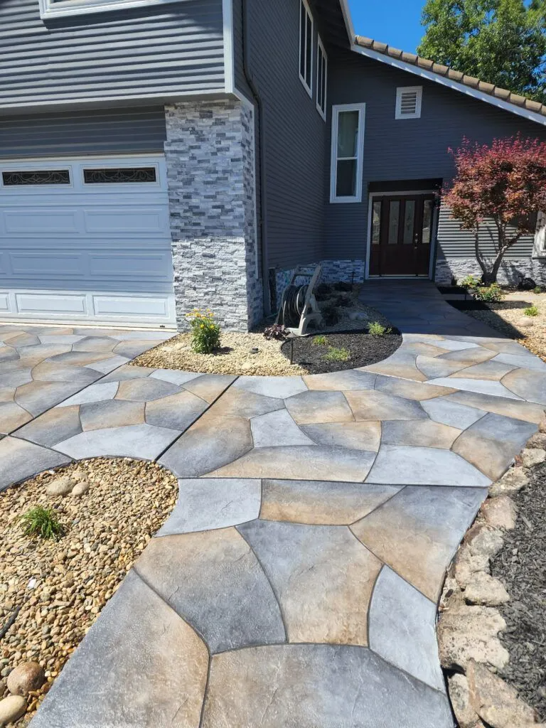 A beautifully designed concrete walkway and driveway with intricate patterns, leading to a house. Trees and plants are visible in the landscaping.