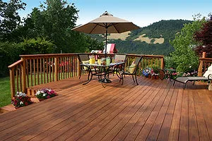 A wooden deck with a patio table, chairs, and an umbrella, surrounded by a wooden railing, set in a scenic outdoor area with hills in the background.