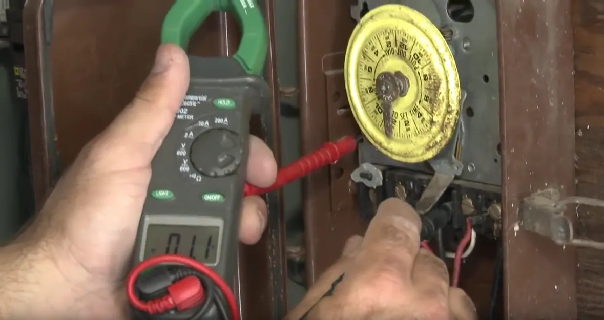 A person's hand using a multimeter to test an electrical connection next to a yellow time switch.