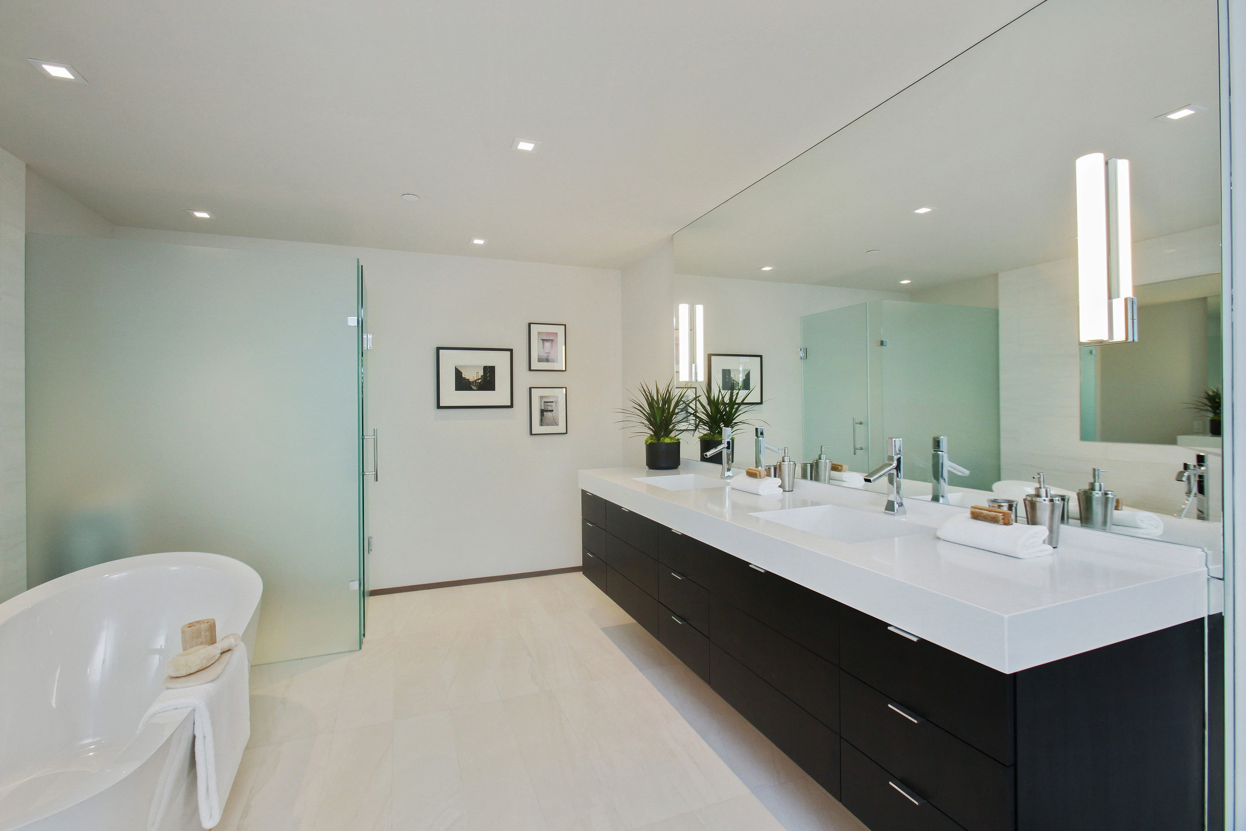 A modern bathroom with a large mirror, double sink vanity, white countertops, dark cabinets, a frosted glass shower door, and a freestanding white bathtub.