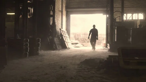 A silhouette of a person walking towards the exit in a dimly lit industrial warehouse, with beams of light streaming in through the opening.