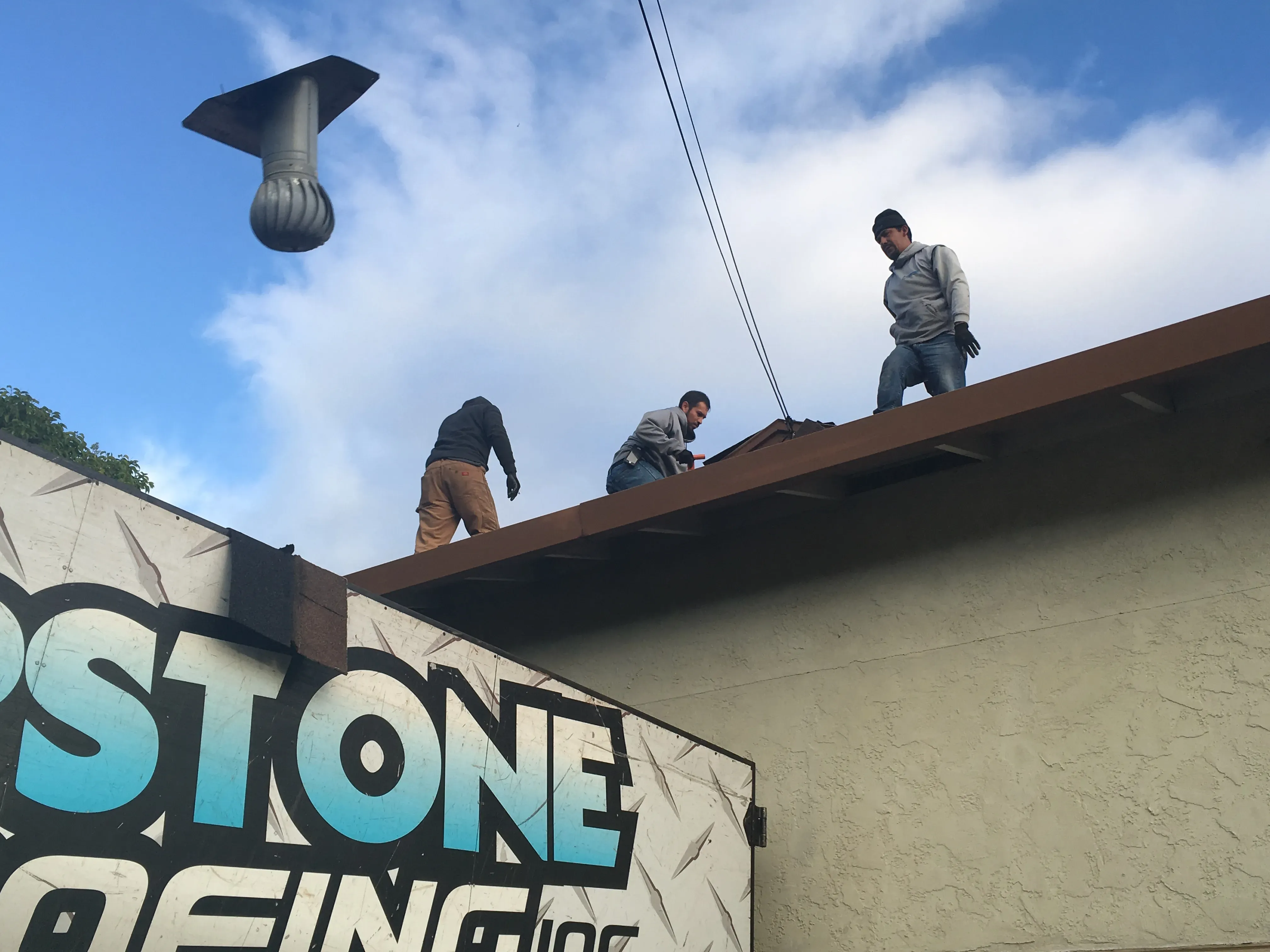 Three workers standing on a roof in Santa Rosa, wearing work attire, with a visible sign saying "Capstone Roofing, Inc." and a vent structure in the sky.