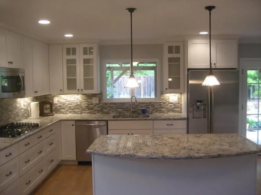 A modern kitchen with white cabinets, granite countertops, and stainless steel appliances, featuring a central island and pendant lighting.