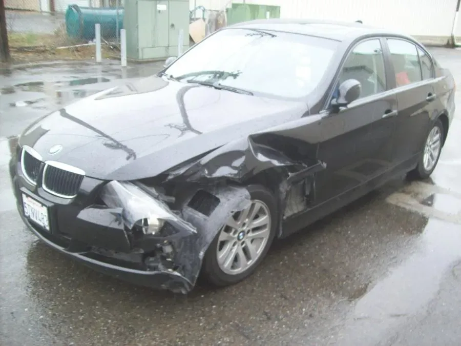 A black car with significant front-end damage, displayed in a parking lot on a rainy day.