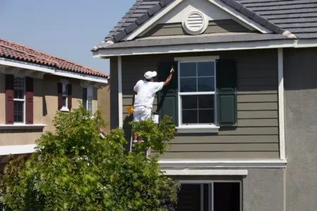A painter from Cartwright Painters LLC on a ladder, painting the shutters of a two-story house in San Jose.