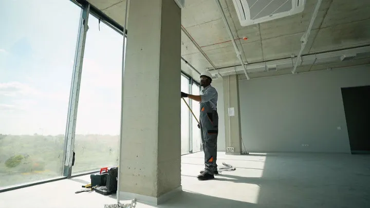 A painter from Cartwright Painters LLC working on a large window frame in an unfinished room with concrete floors and ceilings, showcasing a view of the outdoors in San Jose.
