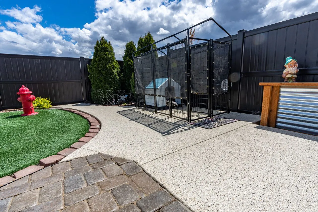 Outdoor area with a stone pathway, artificial grass, a red fire hydrant, a fenced dog area, a decorative gnome, and well-painted surfaces under a cloudy sky.