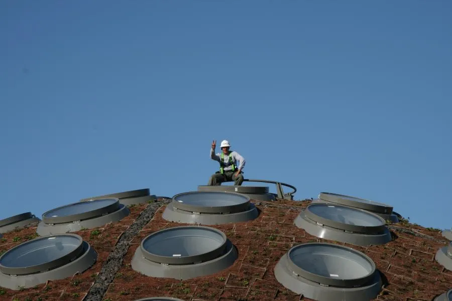 A person in a hard hat and safety gear stands on a sloped roof with circular skylights against a clear blue sky.