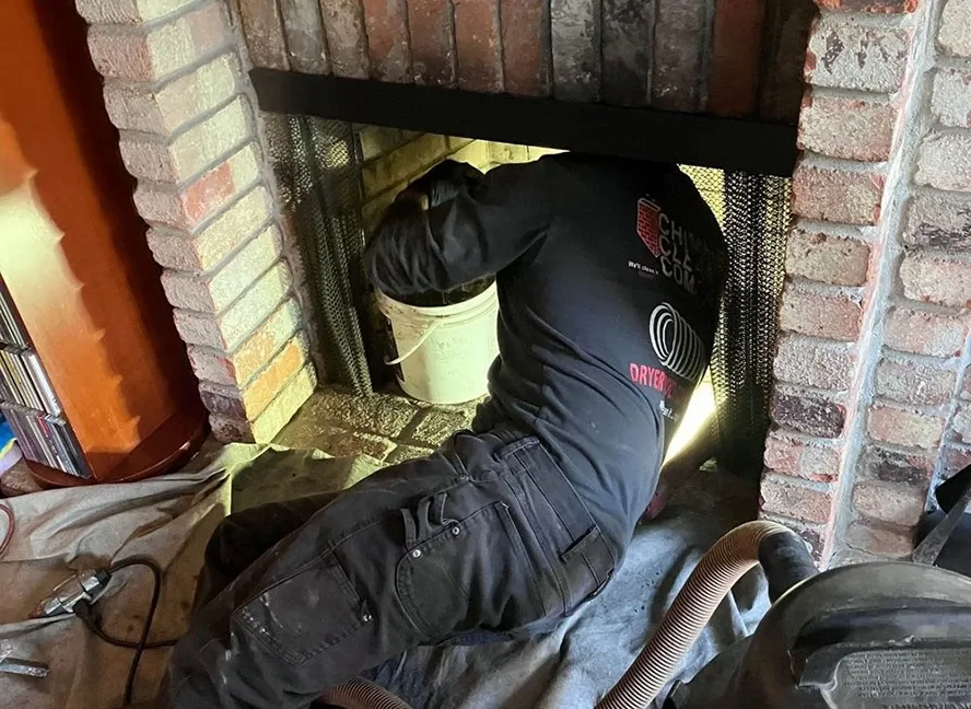 Technician from Chimney Clean Company cleaning a fireplace in San Jose, using a vacuum and a bucket, surrounded by brickwork.