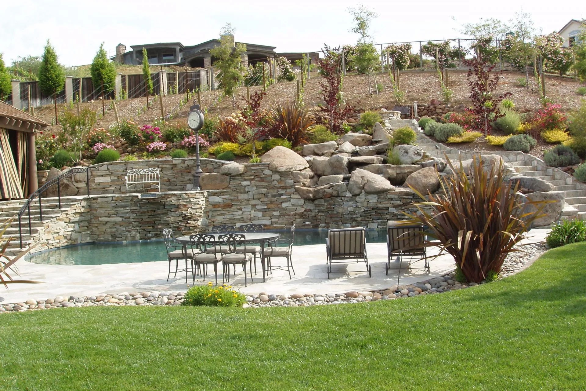 A luxurious backyard pool area designed by Classic Pools in Concord, featuring a stone waterfall, landscaped garden, patio furniture, and surrounding greenery.