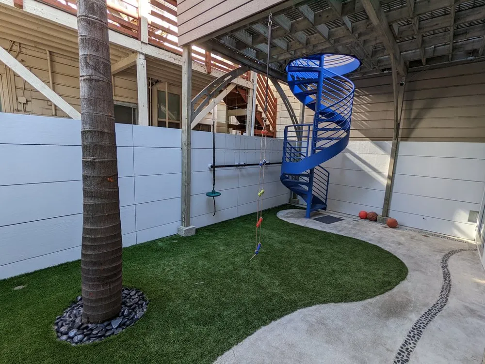 An outdoor area in San Francisco with a blue spiral staircase, artificial grass, a palm tree, and some sports balls. Painted walls provide a clean backdrop.