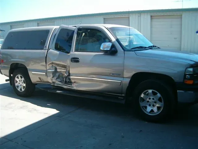 A damaged silver pickup truck with a prominent dent on the driver's side door is parked in front of a garage in Fremont.