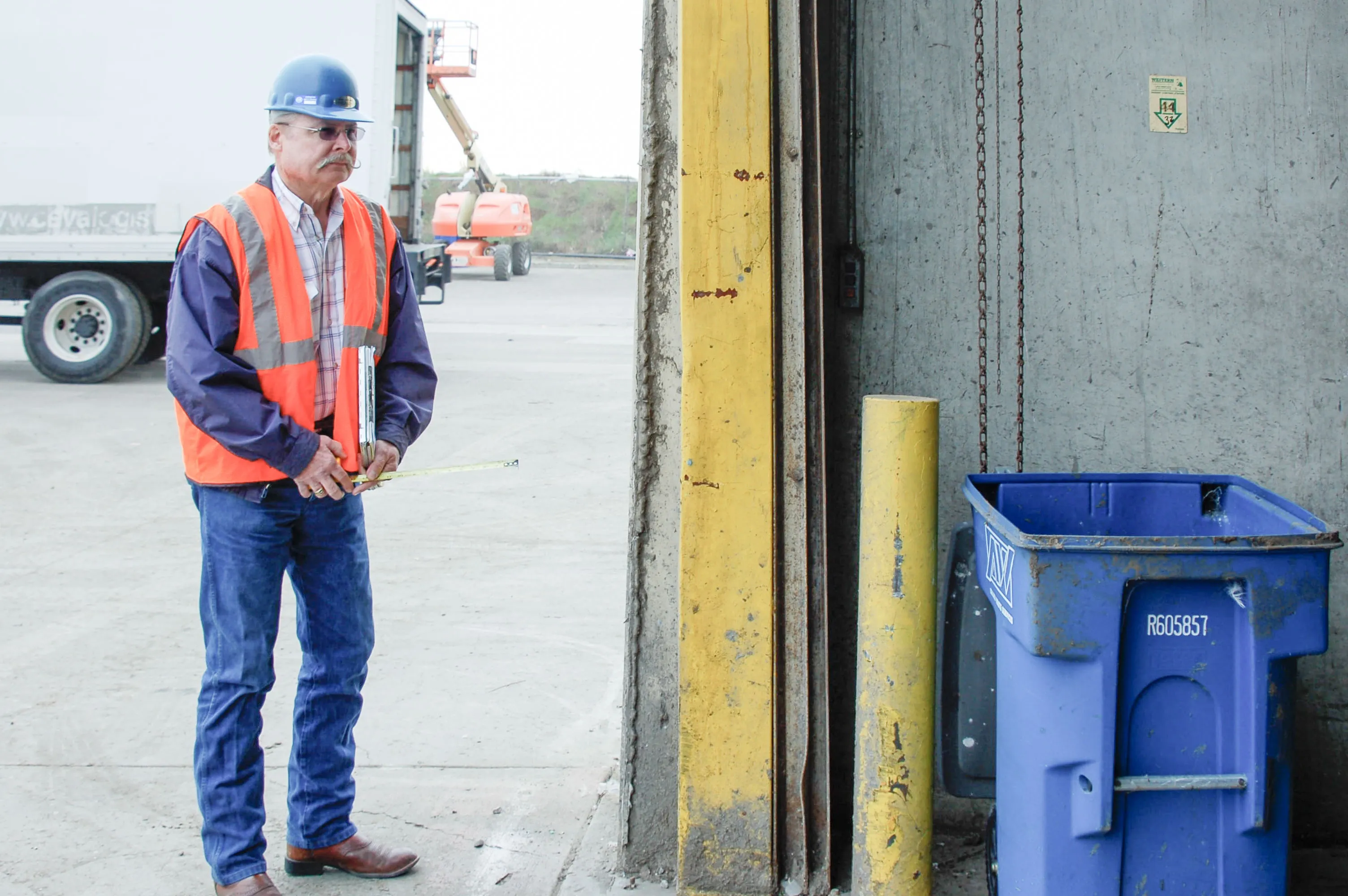 A worker in a blue hard hat and orange safety vest stands next to a blue recycling bin in an industrial area.