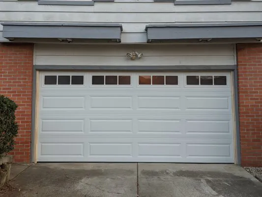 A white garage door with rectangular windows at the top, installed in a brick and white paneled building.