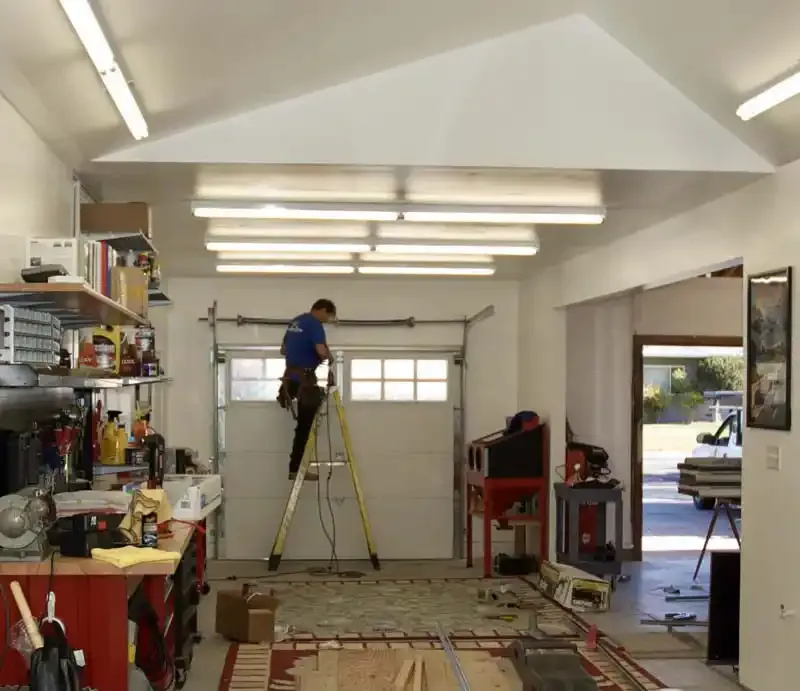 A technician from Contra Costa Door working on a garage door installation in a well-organized workshop in Concord, standing on a ladder with various tools around.