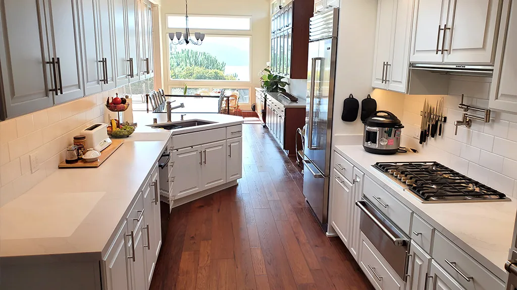 A modern kitchen with white cabinets, a gas stove, a countertop appliance, wooden flooring, and a view of large windows overlooking a scenic landscape, designed by Cook’s Kitchen & Bath, Inc. in Benicia.