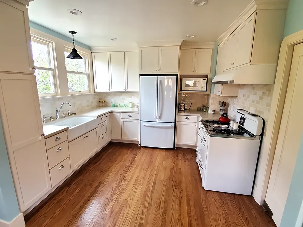 A modern kitchen with white cabinetry, wooden flooring, a farmhouse sink, and stainless steel appliances, designed by Cook's Kitchen & Bath, Inc. in Benicia.