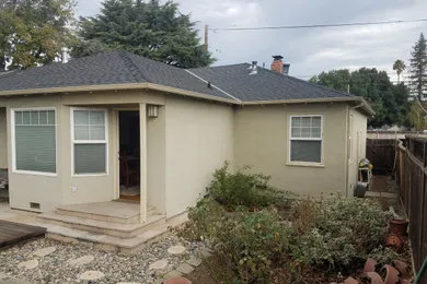 A modest light-colored house with a dark roof, featuring a small front porch and surrounded by a garden, completed by Craft Builders Construction in Morgan Hill.