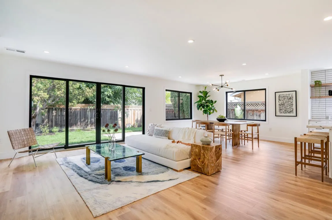 A modern open-plan living room and dining area featuring large glass doors, wood floors, a white sofa, a glass coffee table, and a dining table, designed by Craftsmen's Guild, Inc. in Cupertino.