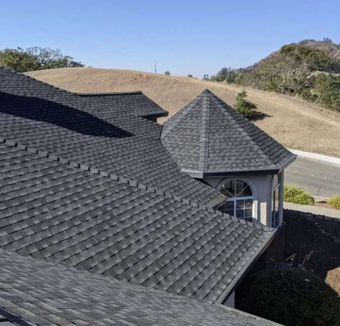 A detailed view of a well-constructed shingle roof with a turret, showcasing the craftsmanship of Crandall Roofing Inc. in Santa Rosa against a backdrop of rolling hills and a clear sky.