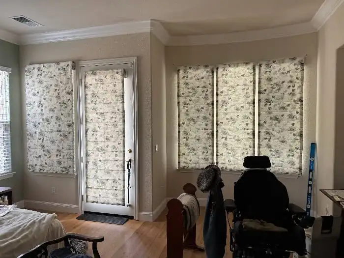 A room featuring light-colored floral-patterned window coverings, with a wheelchair, furniture, and wooden flooring, showcasing interior design by Creative Window Fashions in Rohnert Park.