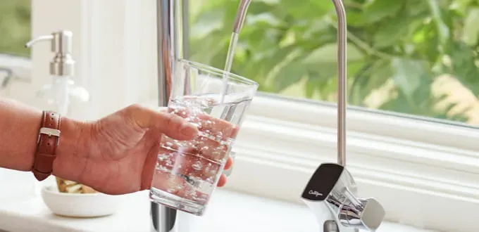 A person fills a glass of water from a Culligan-branded kitchen faucet, with a green plant visible outside the window.