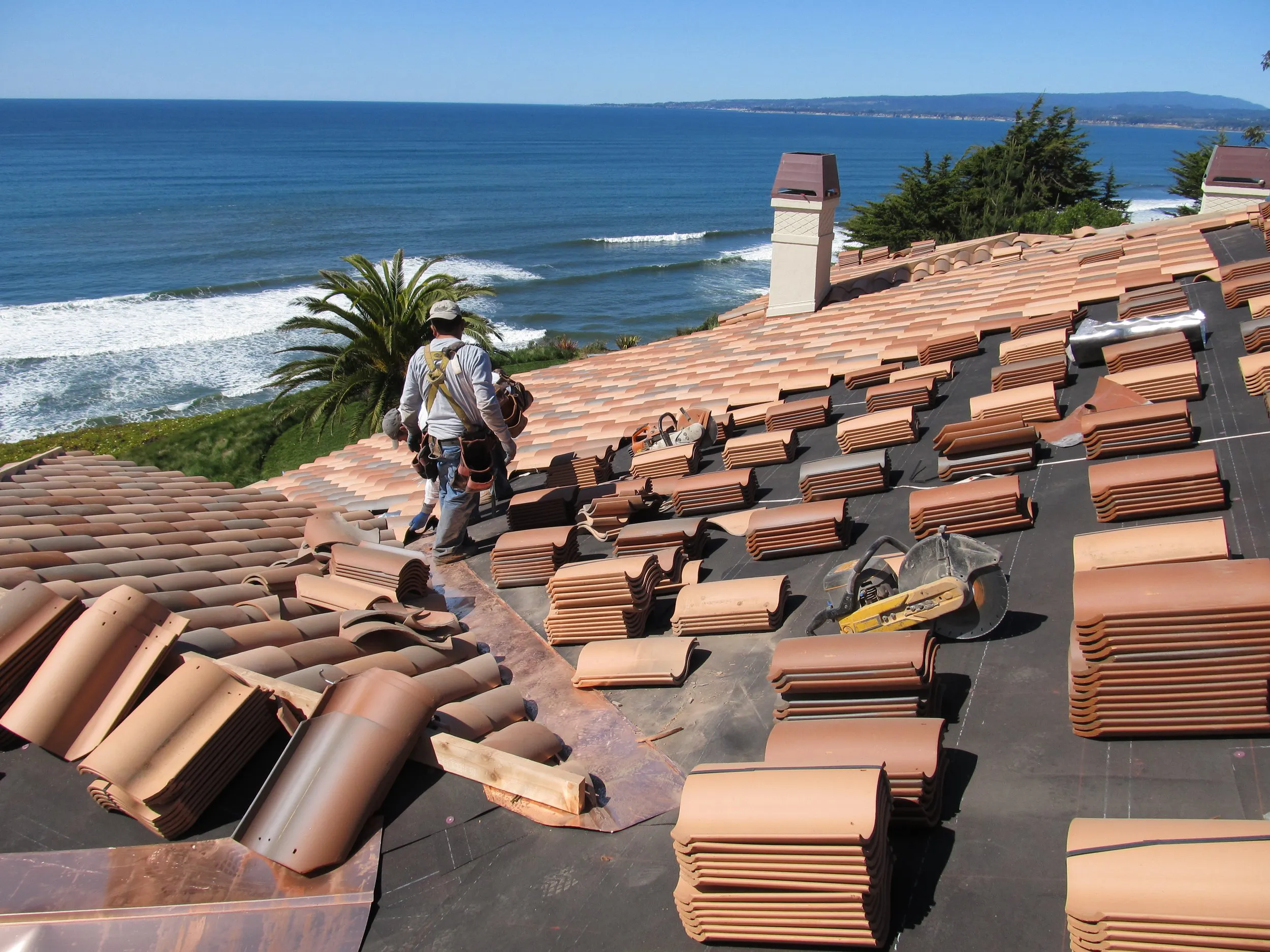 A person works on a tiled roof overlooking the ocean, with stacks of roofing tiles and tools, representing the roofing services of Daddario Roofing Company, Inc. in Capitola.