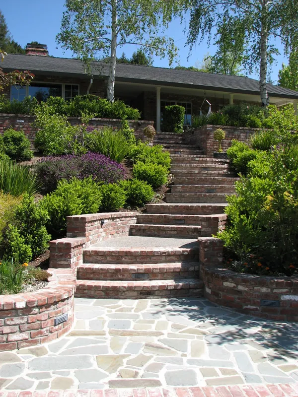 A beautifully landscaped garden with a brick pathway leading up to a house, surrounded by lush greenery and shrubs, designed by Dana Lund Landscaping in San Ramon.