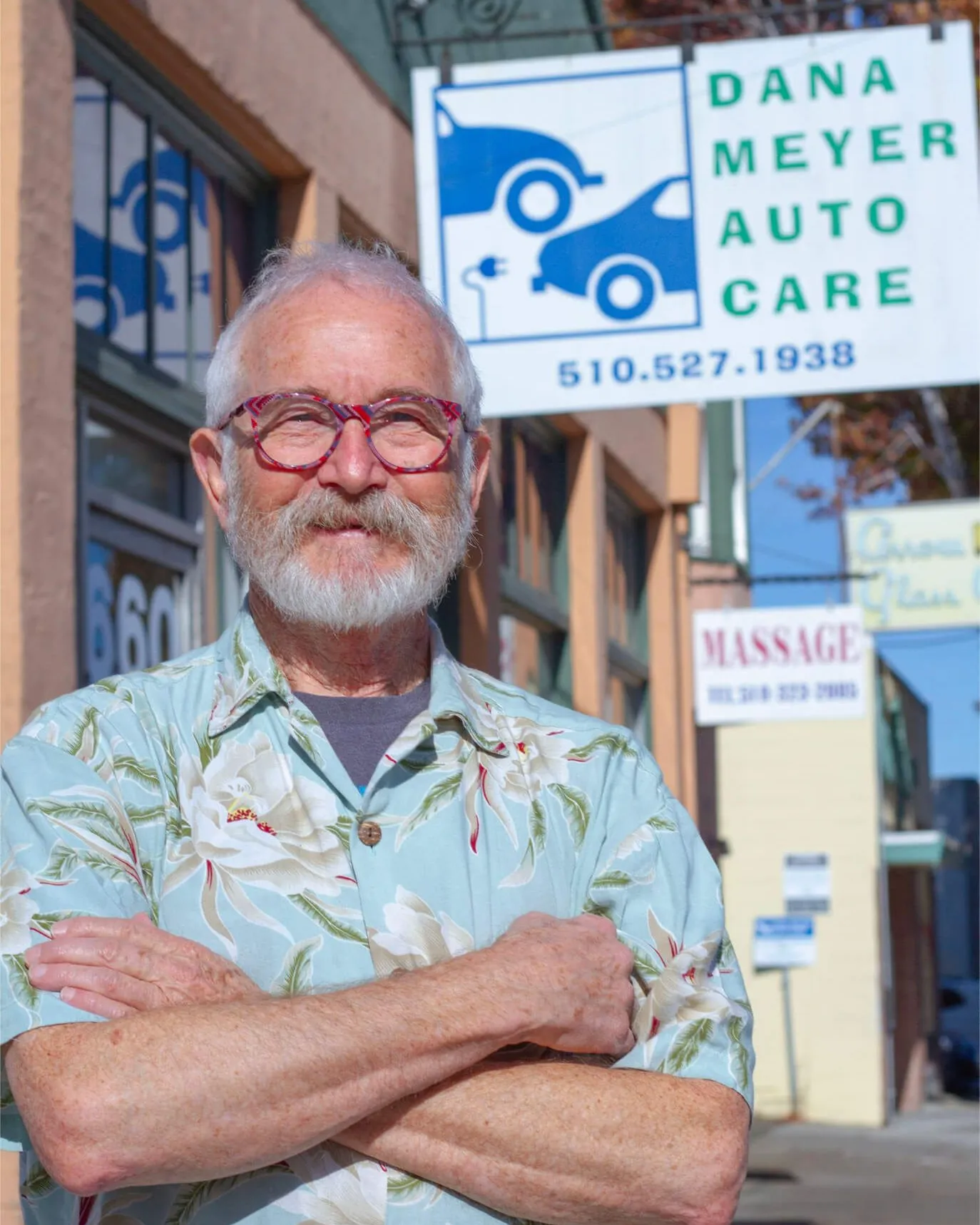 A person with glasses and a floral shirt stands outside DanaMeyerAutoCare in Albany, with the business sign visible in the background.