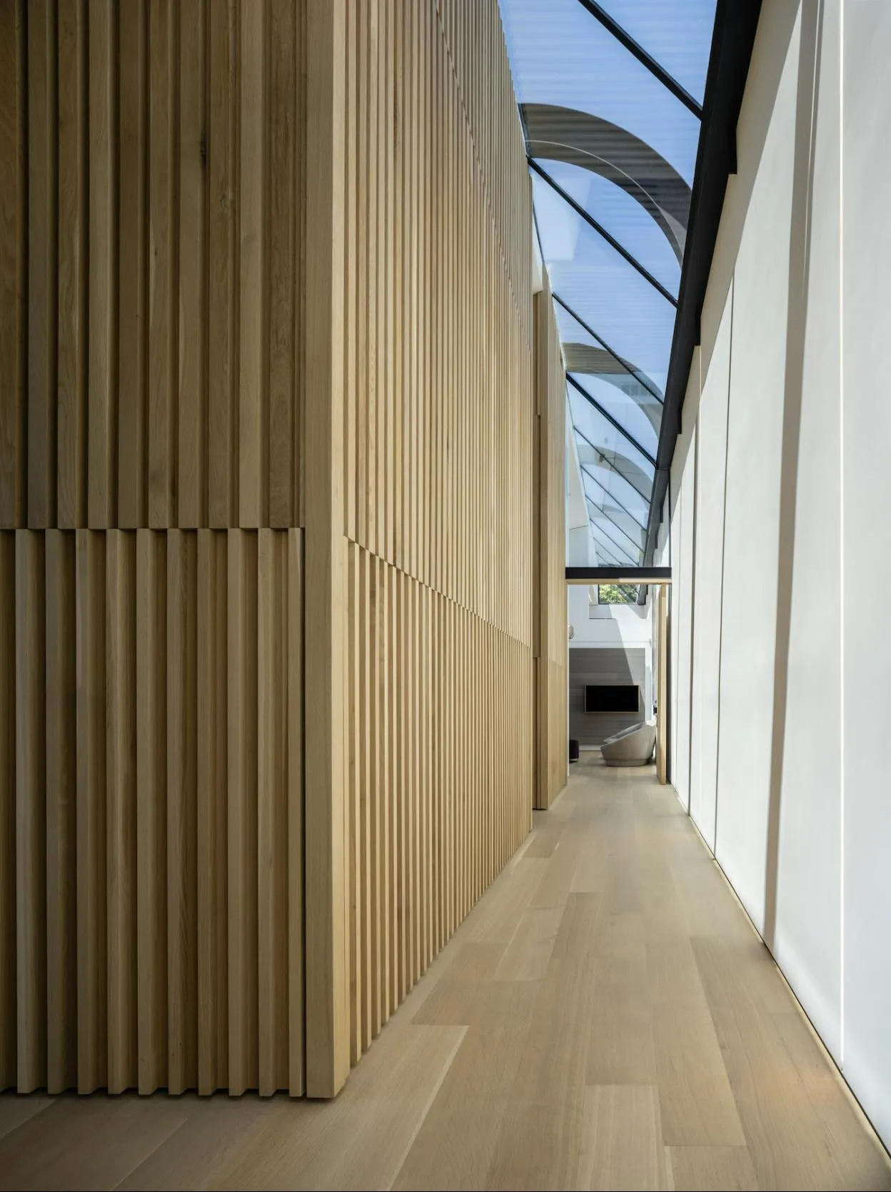 A sleek and modern hallway featuring vertically paneled hardwood walls and polished wood flooring, illuminated by natural light through a skylight.