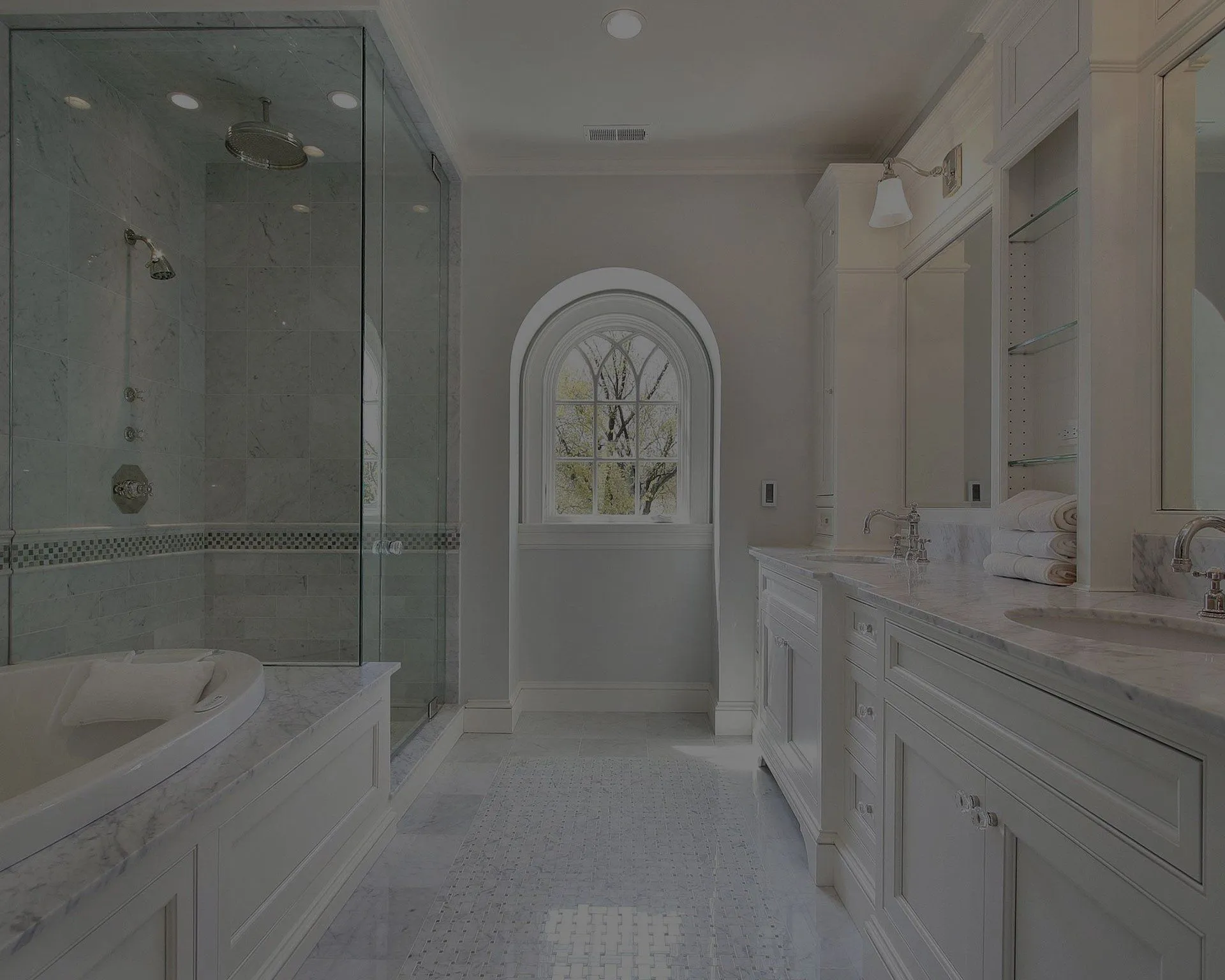 A modern bathroom featuring a glass-enclosed shower, a freestanding bathtub, double sinks on a marble countertop, white cabinetry, and an arched window.