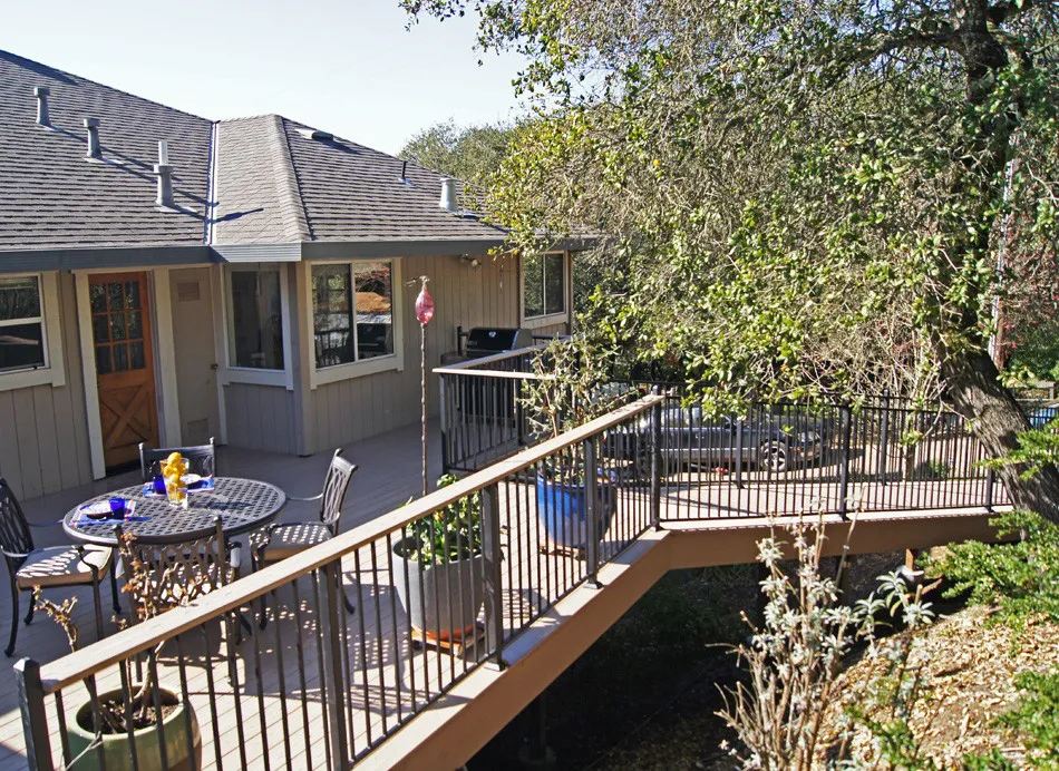 A spacious outdoor deck with seating and a table, surrounded by trees, attached to a house, designed by Deckmaster Fine Decks, Inc. in Sebastopol.
